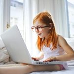 selective focus photo of young girl in glasses sitting in bed while using a laptop